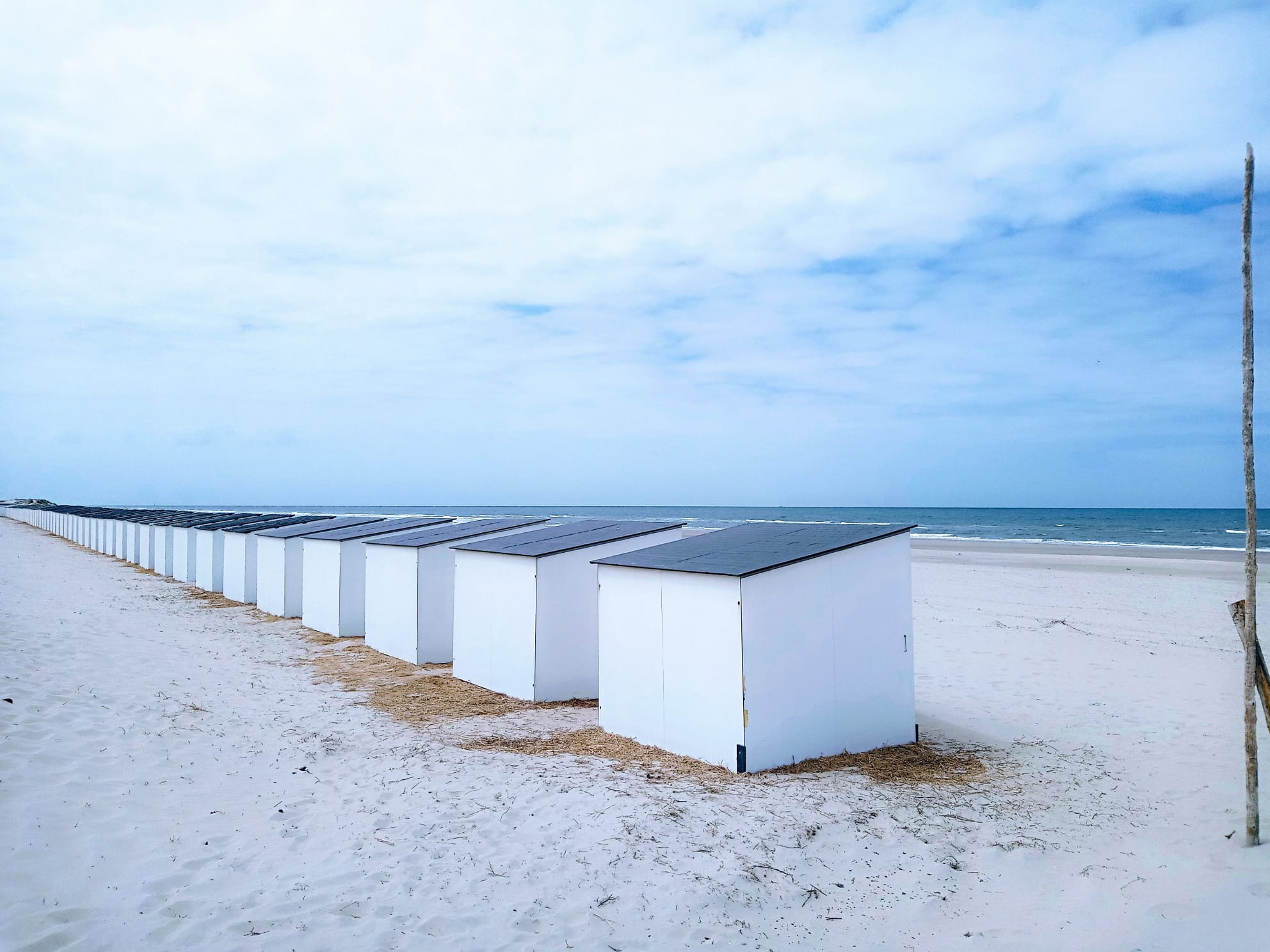 Reihe weißer Strandhäuschen mit dunklen Dächern am leeren Sandstrand, im Hintergrund das Meer unter bewölktem Himmel.